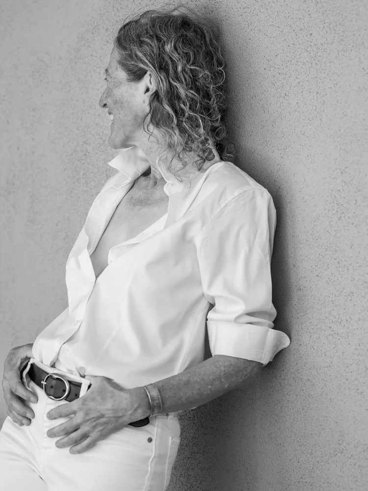 A woman laughing in profile in a white shirt against a sunlit wall, photographed by Deborah Jaffe